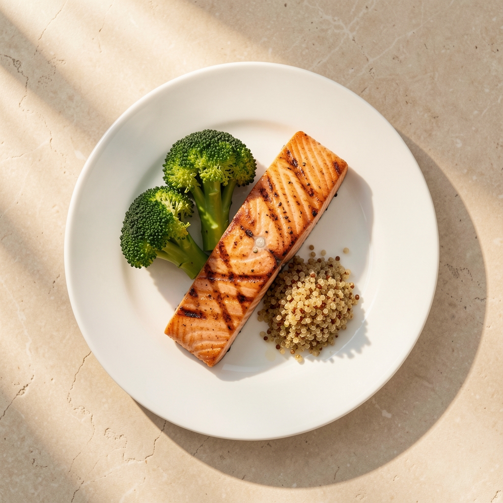 Overhead view of a white plate with grilled salmon, steamed broccoli, and a small portion of quinoa on a pale stone surface with warm natural light